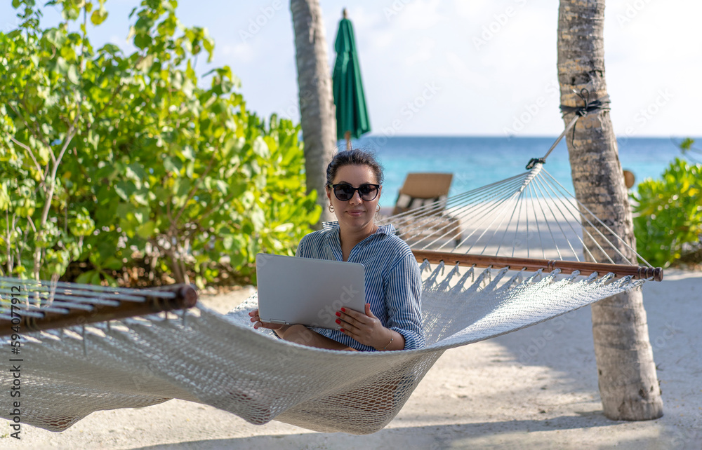 A young woman works on a laptop in a hammock on the beach in the Maldives. The concept of remote work and nomadism