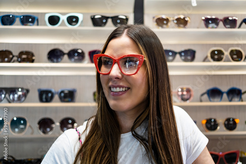 A beautiful young woman stands inside an optician posing with Graduates glasses in front of a rack of glasses. Concept of sale of glasses. Choice of glasses. Glasses model. photo
