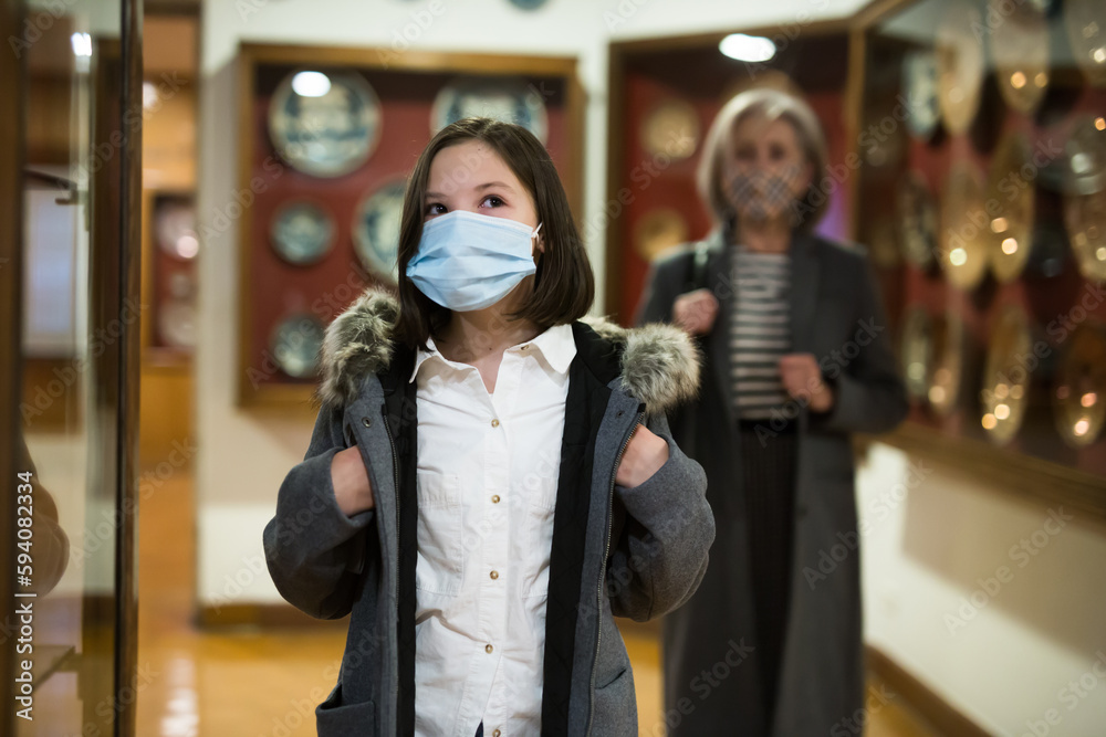 Cute interested preteen girl wearing protective face mask exploring artworks in modern museum of applied arts. Forced precautions in pandemic