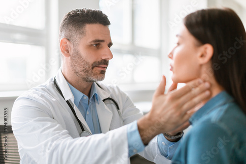 Friendly middle aged male doctor checking sore throat or thyroid glands, touching neck of young female patient