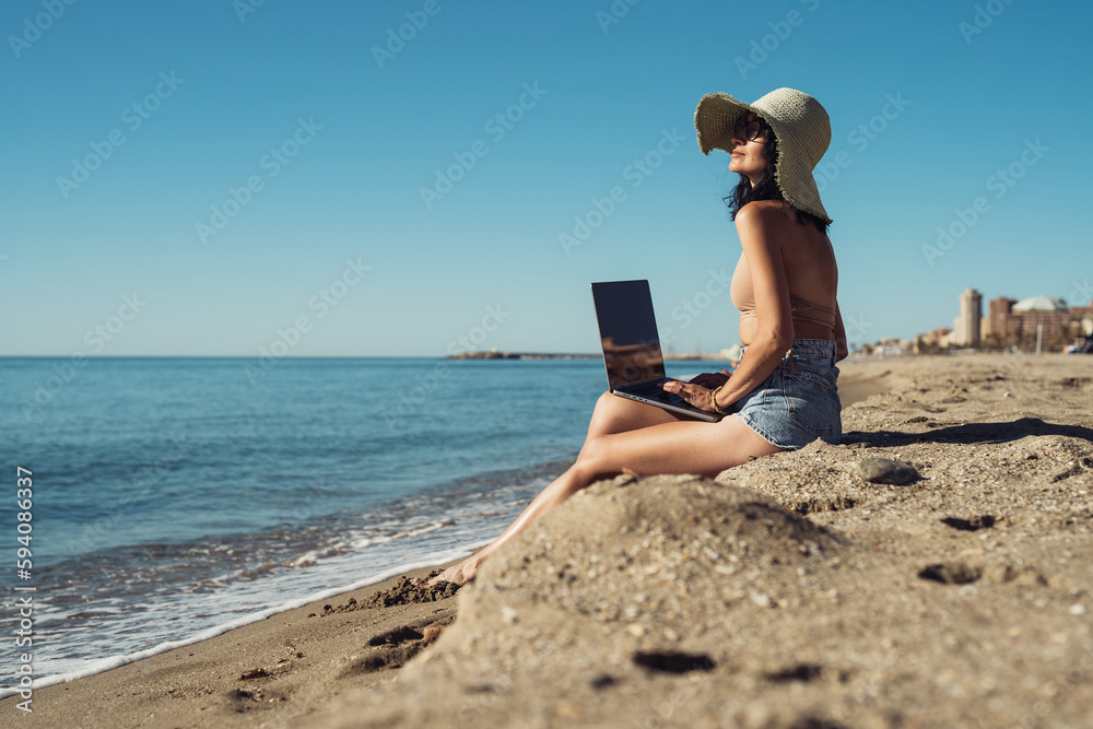 young beautiful girl freelancer with a laptop works on the seashore sitting on the sand
