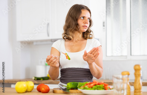 Portrait of displeased woman in nightie eating salad in kitchen at home. Dissatisfied woman having healthy dinner.