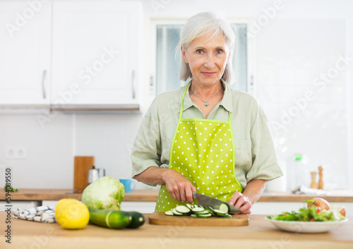 Positive old woman in apron cooking salad, standing at kitchen table and slicing vegetables.