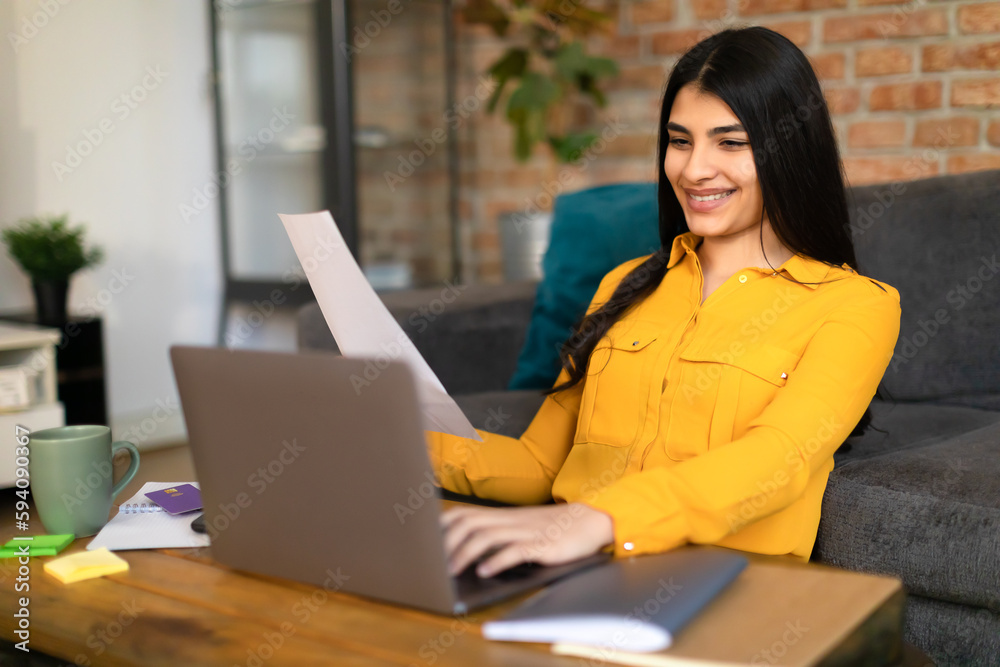 Happy spanish female student sitting at table with laptop and notepads, holding sheet of paper, writing coursework Happy spanish female student sitting at table with laptop and notepads, holding sheet of paper, writing coursework