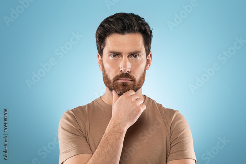Headshot of attractive focused middle aged man looking at camera and touching unshaven chin, posing on blue background