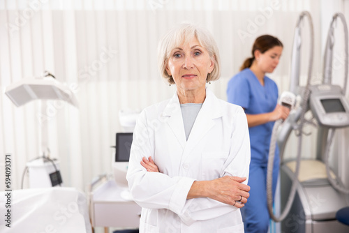 Elderly woman cosmetologist looking at camera while her assistant nurse adjusting vacuum massage machine in clinic of aesthetic medicine