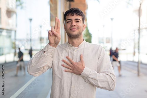 young handsome man looking happy, confident and trustworthy, smiling and showing victory sign, with a positive attitude