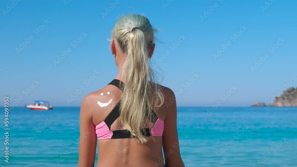 Emotional sunbathing with sunblock. A little girl sitting on the beach with smiling emoticon on her shoulder. A concept of taking care on skin during traveling under hot summer sun.