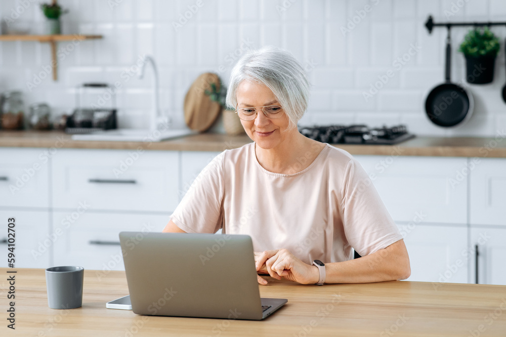 Positive modern stylish gorgeous caucasian mature gray haired woman, sits at home in the kitchen, uses laptop for remote work, online shopping, read news, looks satisfied, smiles Positive modern stylish gorgeous caucasian mature gray haired woman, sits at home in the kitchen, uses laptop for remote work, online shopping, read news, looks satisfied, smiles