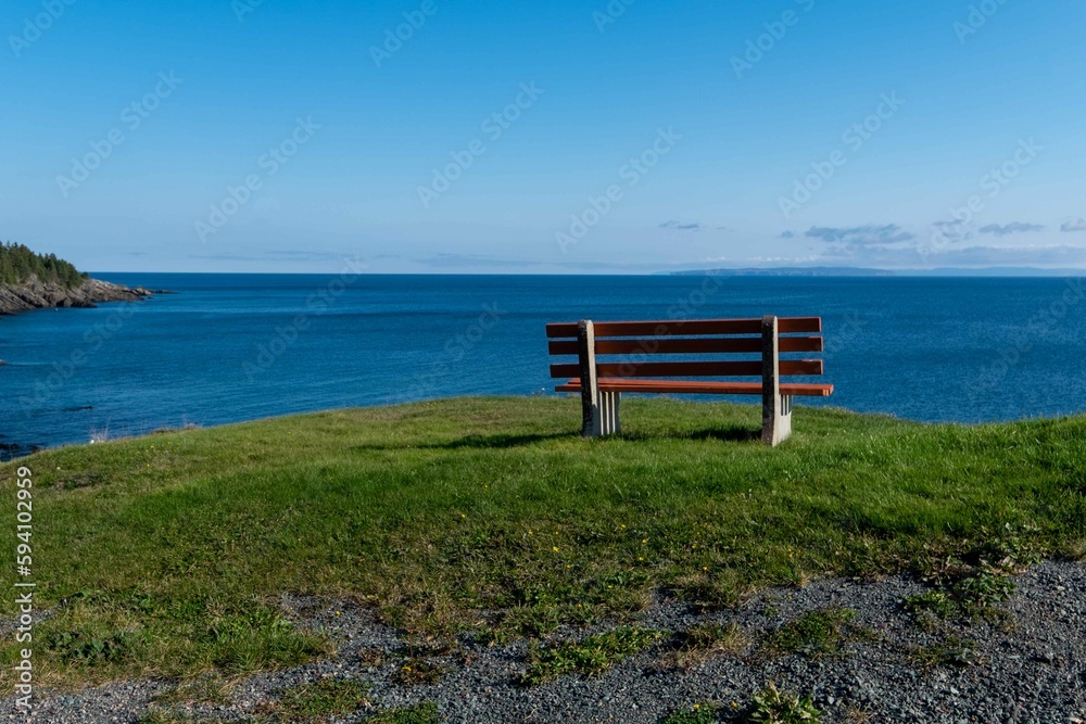 A brown wooden bench with concrete bases on a hill overlooking the blue ocean. The coastline is rocky with jagged rocks. The coast is covered in green evergreen trees. The water is calm blue.