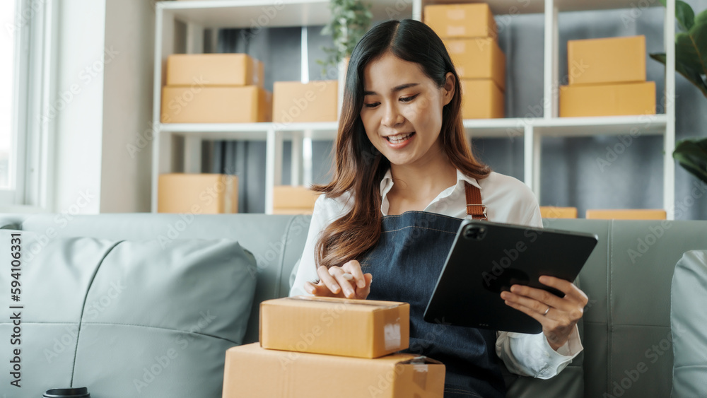 Using tablet computer, Young pretty Asian female influencer small businesses SME owners female entrepreneurs working on receipt box and check online orders, sitting on sofa of home online business. Using tablet computer, Young pretty Asian female influencer small businesses SME owners female entrepreneurs working on receipt box and check online orders, sitting on sofa of home online business.