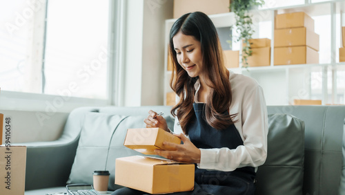 Writing clients address, Young pretty Asian female influencer small businesses SME owners female entrepreneurs working on receipt box and check online orders, sitting on sofa of home online business. © makibestphoto