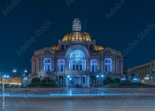 Palacio de Bellas Artes en la noche, cielo azul despejado. Fotografía de larga exposición, rastros de luces de carros en la acera. 