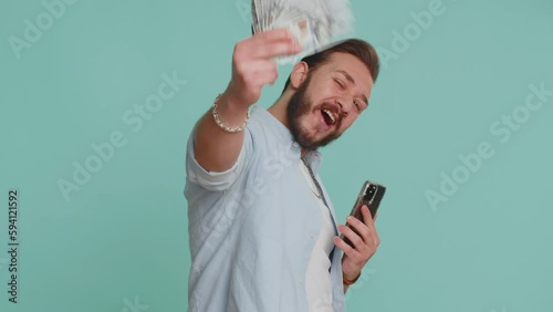 Lebanese man looking smartphone display sincerely rejoicing win, receiving money dollar cash banknotes, success lottery jackpot luck, income salary wage. Arabian guy isolated on studio blue background photo