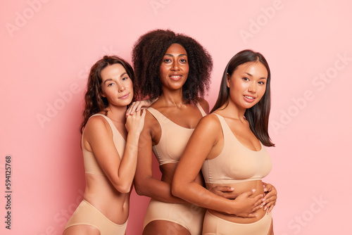 Women of different races dressed comfortable beige underwear clothig stand half-turned inside the studio, one puts her hand on shoulder of her friend next to, another puts her hand on waist, modesty