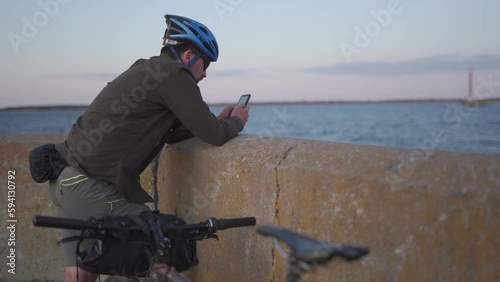 Cycling trip to sea at sunset. Male cyclist is resting on embankment by sea and uses smartphone to build route and navigate GPS. Male with bike on seashore surfing internet on a smart phone. photo