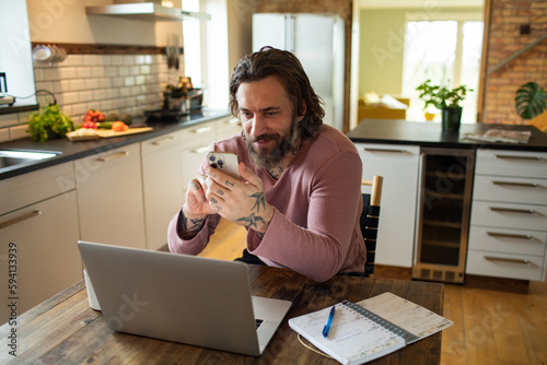 Young adult man using a smart phone in the kitchen while working from home