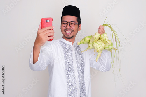 Moslem Asian man smiling happy when calling his family while showing rhobus shaped rice cake photo