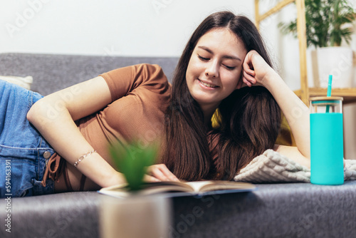 Teen girl lying on the couch and reads a book. Modern teenager at home. Teen literature concept.