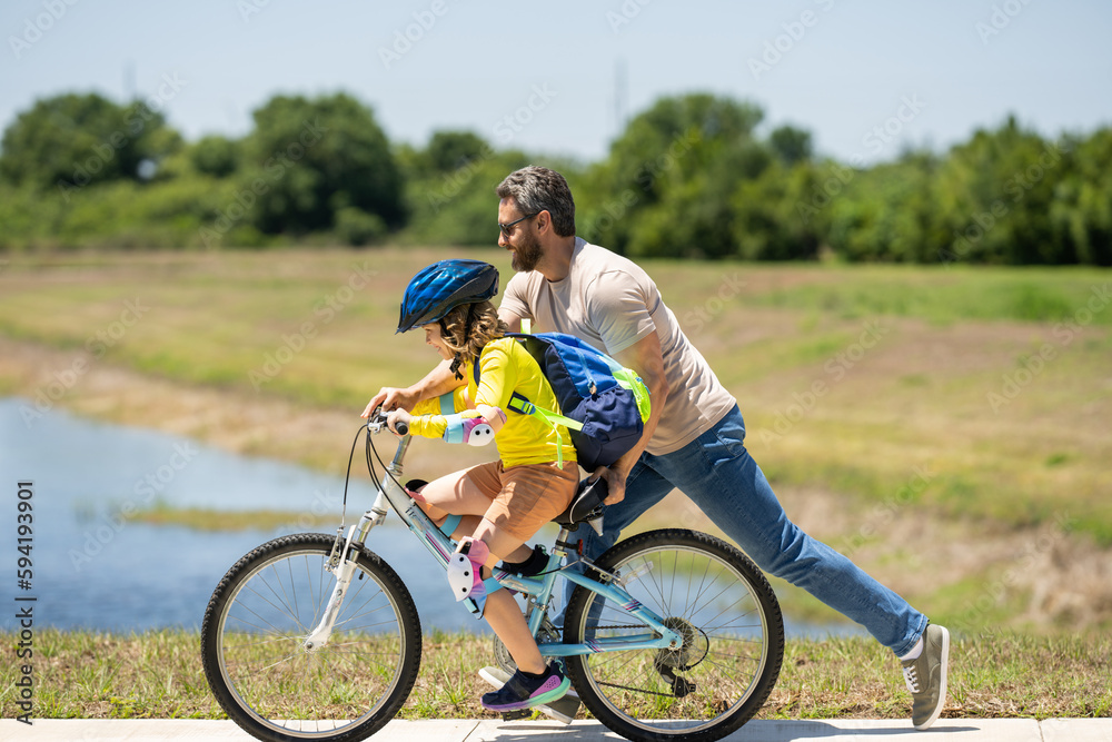 Fototapeta premium Happy Fathers day. Father and son in bike helmet for learning to ride bicycle at park. Father helping son cycling. Father and son on the bicycle on summer day. Kid son trying to ride bike with father.