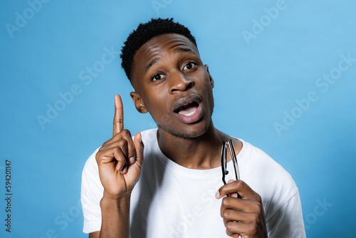 I have idea. Curious attractibe african american schooler student with glasses in hands opening mouth showing index finger at camera over blue background in studio. photo