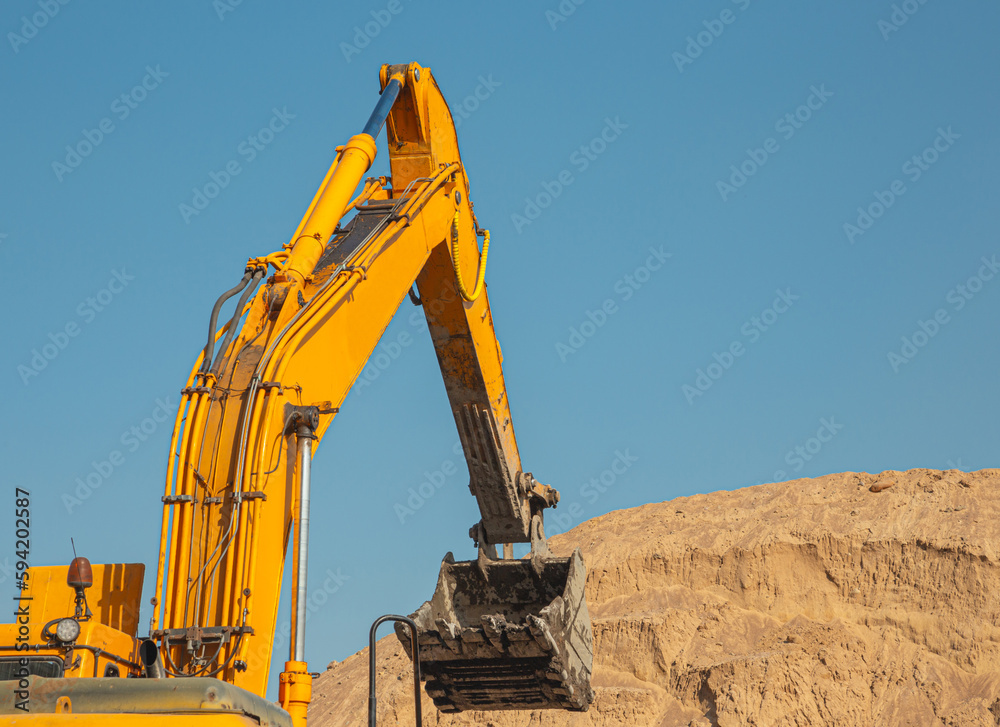 An orange excavator fills up the earth in the body of a construction dump truck. Excavator with bucket and dump truck, while working on the background of the blue sky close-up. An orange excavator fills up the earth in the body of a construction dump truck. Excavator with bucket and dump truck, while working on the background of the blue sky close-up.
