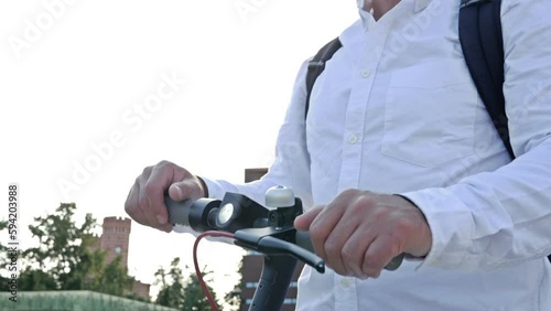 Man in a white shirt and sunglasses stands with an electric scooter in the middle of a city square and admires the architecture.