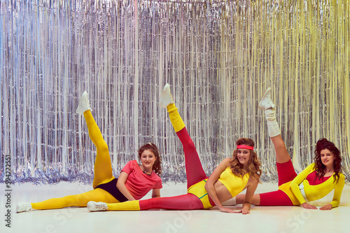 Fototapeta Naklejka Na Ścianę i Meble -  Three stylish young girls in colorful sportswear, bodysuits and leggings training, doing stretching and aerobics exercises. Concept of retro fitness, sport, 80s fashion, beauty, health