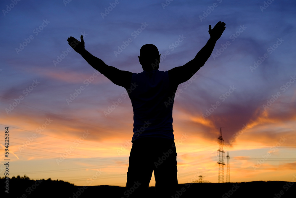 black silhouette of man in delight raises her hands at sunset in a field with silhouette of hills, mountains and colored sky in background, concept of happiness, unlimited freedom