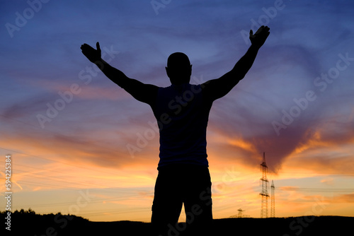 black silhouette of man in delight raises her hands at sunset in a field with silhouette of hills, mountains and colored sky in background, concept of happiness, unlimited freedom