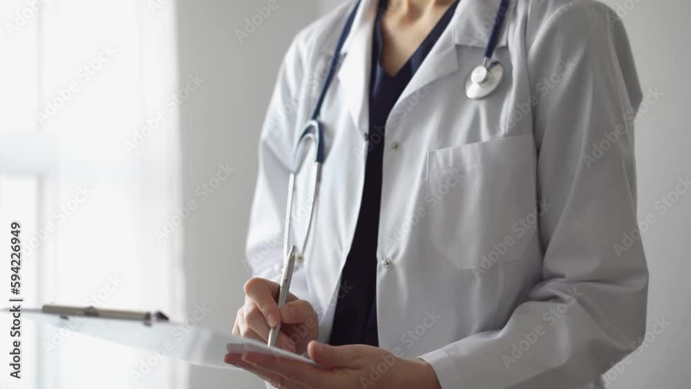 Doctor woman at work using a white clipboard while writing medical notes. An unknown physician standing near the panorama window in hospital. Medicine Doctor woman at work using a white clipboard while writing medical notes. An unknown physician standing near the panorama window in hospital. Medicine