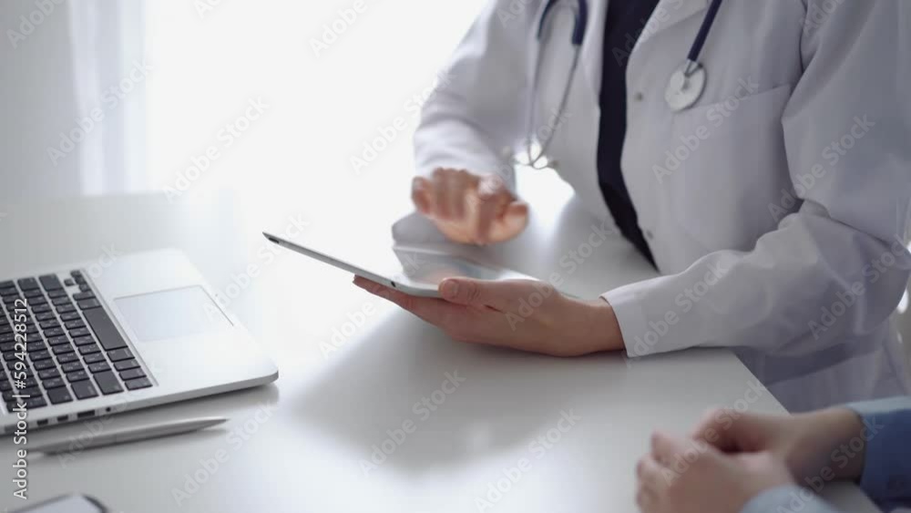 Doctor and patient sitting at the white desk near flair window in clinic. Unknown female physician wearing a white coat uses tablet computer for filling up medical record form. Medicine concept