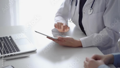 Doctor and patient sitting at the white desk near flair window in clinic. Unknown female physician wearing a white coat uses tablet computer for filling up medical record form. Medicine concept photo