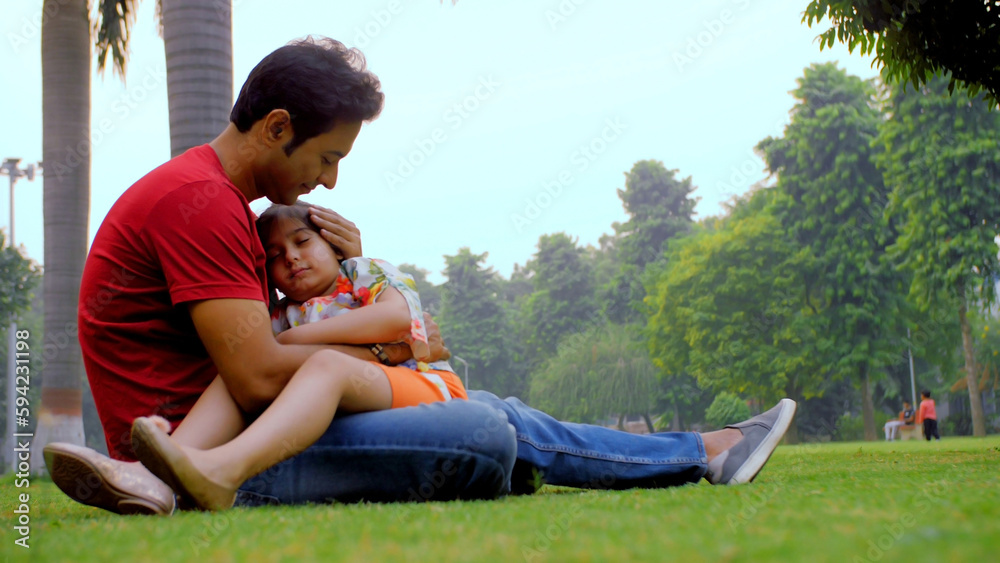 Loving father cuddling his tired and sleepy child sitting outdoors in the park. Adorable little daughter sleeping on her dad's lap in a garden - father's love / family bonding lifestyle concept