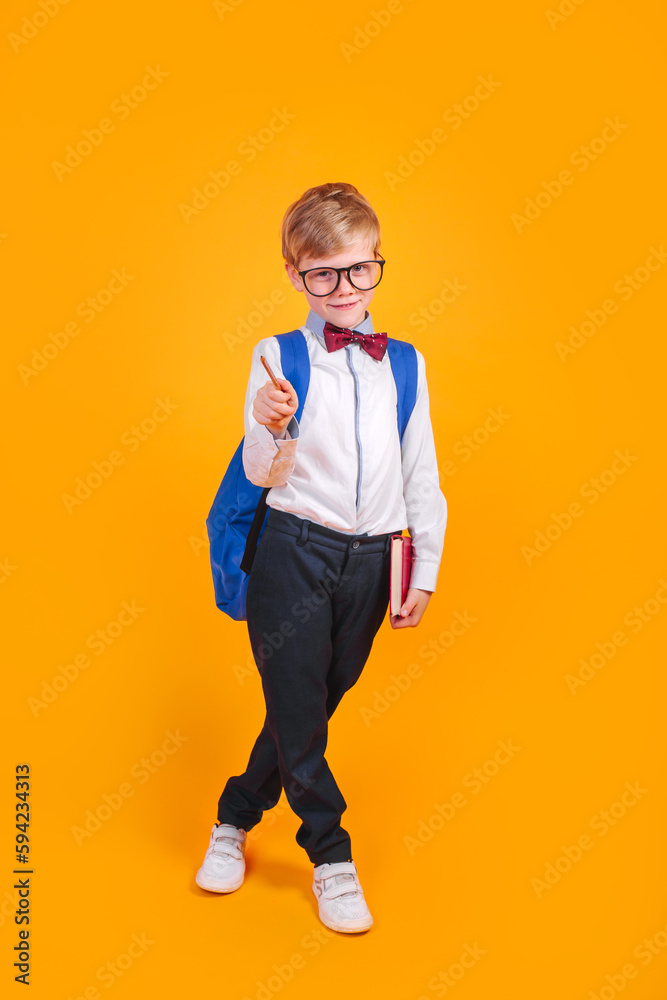 Fototapeta premium Little school boy in eyeglasses and bowtie with backpack holding books and standing on orange background