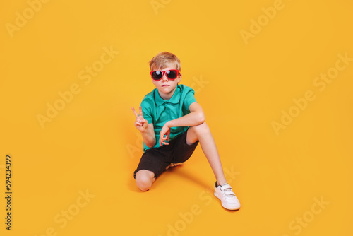 Confident trendy boy in summer clothes and sunglasses sitting on yellow background