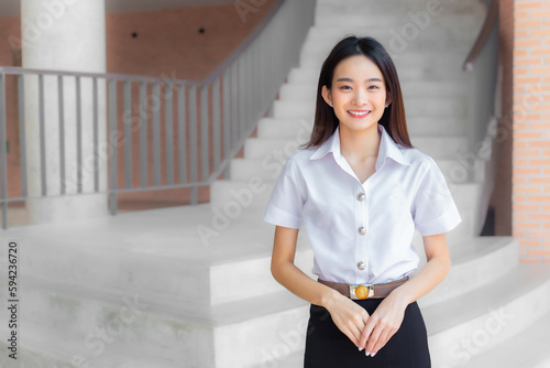 Beautiful Asian young woman student in Thai student uniform is stand under the school building smiling and looking at camera in university background.