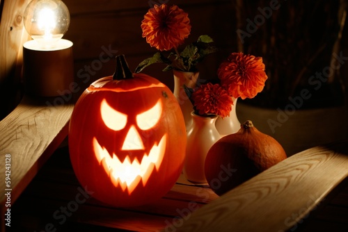 Close-up shot of a carved spooky illuminated pumpkin and flowers in a vase on a wooden table