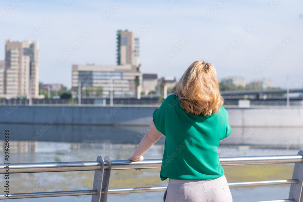 A view from the back of a girl in a green blouse standing on the riverbank against the background of a blurred city.