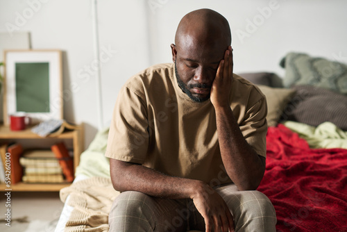 African American man sitting on bed in depression with his eyes closed and having headache