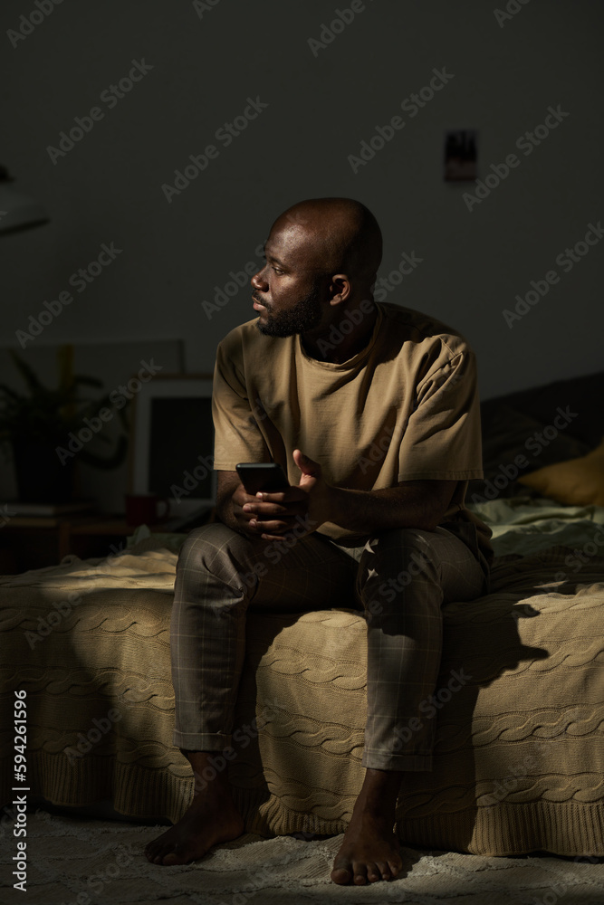 African American man sitting on bed with mobile phone and thinking about something