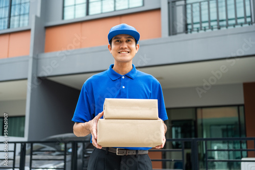 Asian delivery man with parcel in hand of blue uniform sending parcel to customer front of the house from shopping online with good service. Courier man send a package to destination.