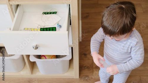 Toddler baby opened the cabinet drawer with pills and medicine. Child boy holding a pack of pills in the home living room. Kid aged about two years (age one year nine months)