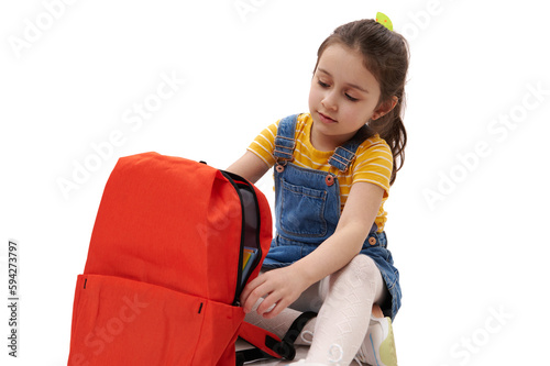 Close-up portrait of 5-6 years old Caucasian adorable little child, smart lovely schoolgirl in denim sundress, posing with orange backpack, isolated over white background. Education and school concept