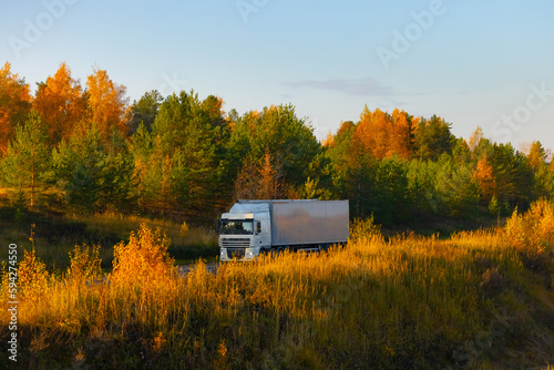 A truck of an unknown brand is driving on a road in an autumnal landscape bathed in the light of the setting sun. The autumn colors, the romance of a long-haul trucker, and the delivery goods on land.