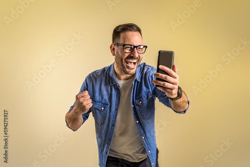 Successful male freelancer reading good news over mobile phone and screaming cheerfully against beige background. Young man dressed in denim shirt pumping fist while messaging on cellphone photo