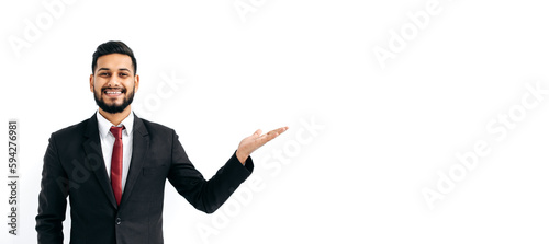 Copy-space for your presentation. Panoramic photo of positive elegant arabian or indian business man, stand on isolated white background, looks at camera, pointing hand to empty space, smiles happily