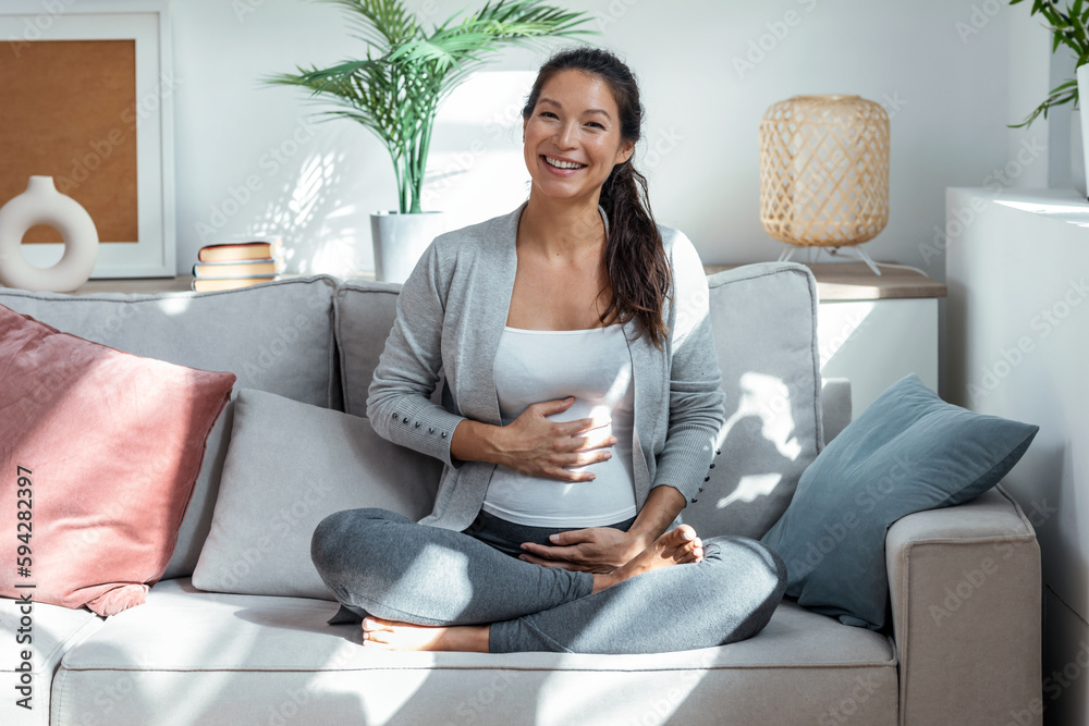 Beautiful pregnant woman touching her belly sitting in lotus position on couch in living room at home. Beautiful pregnant woman touching her belly sitting in lotus position on couch in living room at home.