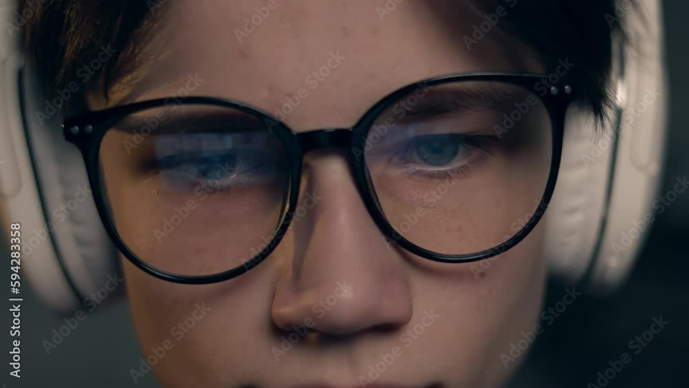 Young gamer man in glasses with screen reflection and headphones streaming live action game while explaining gameplay to subscribers. Gaming, esports, internet and virtual games at dark room. Young gamer man in glasses with screen reflection and headphones streaming live action game while explaining gameplay to subscribers. Gaming, esports, internet and virtual games at dark room.
