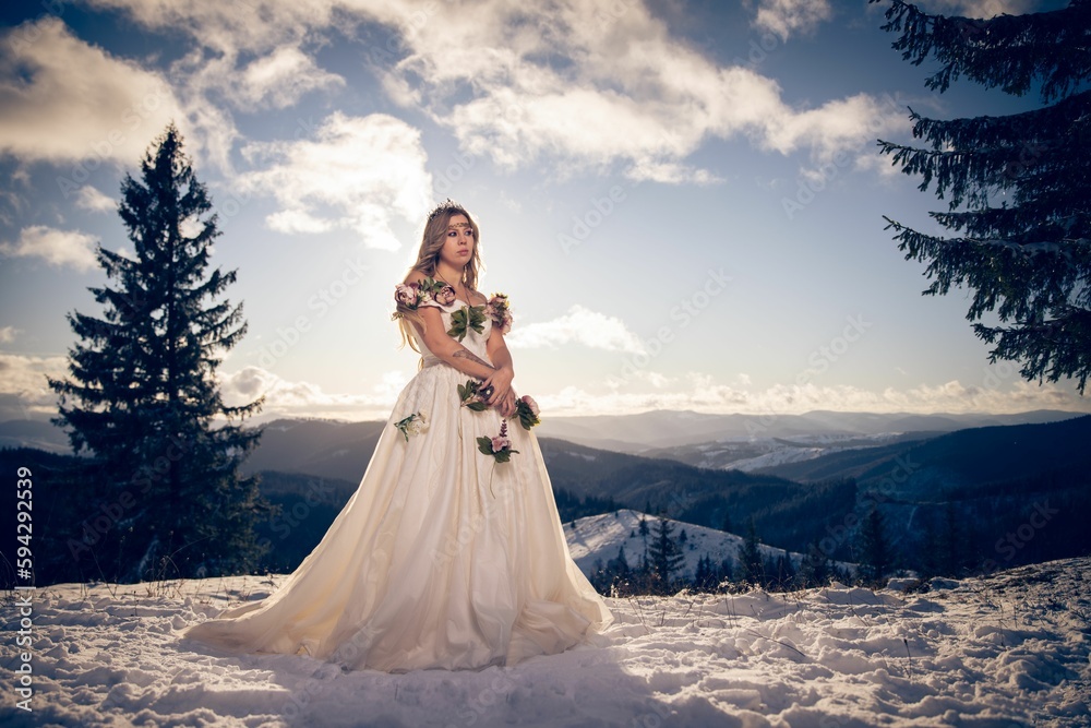 Beautiful young female in a mystical wedding dress with pink flowers posing on a snowy mountain